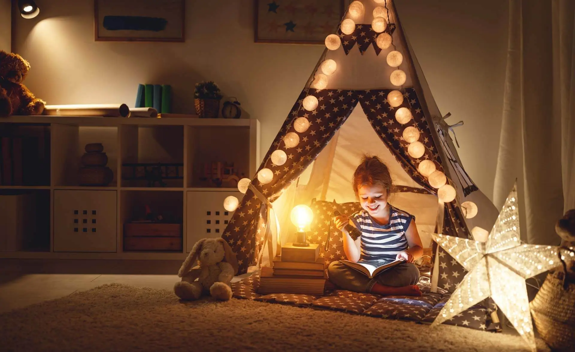 Ein kleines Mädchen sitzt in seinem Kinderzimmer und spielt in einem kleinen Zelt, welches mit einer Lichterkette dekoriert ist.